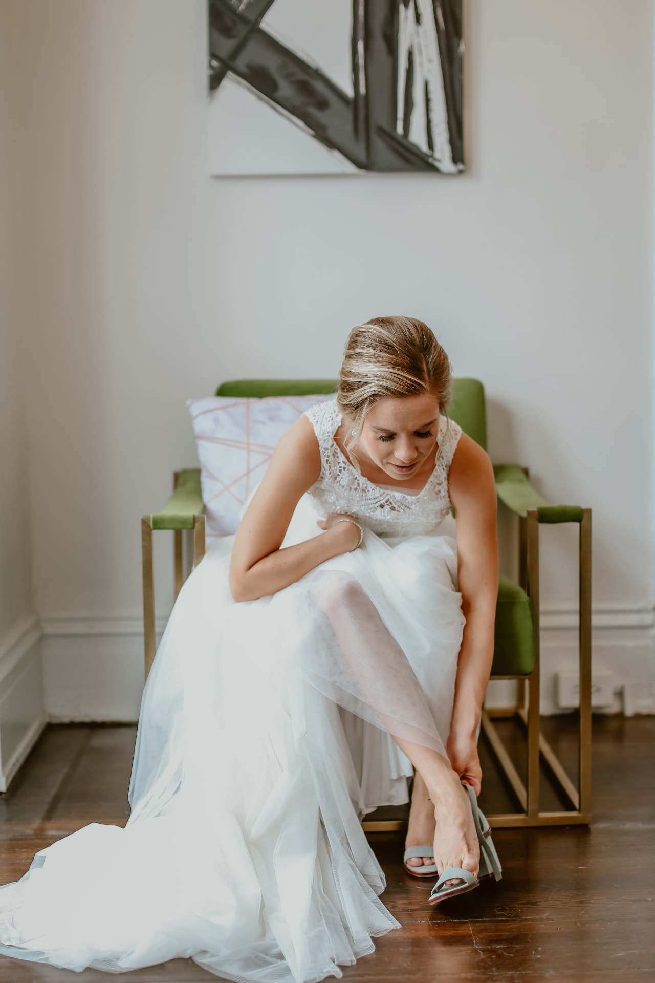 Bride in a lace gown fastening her shoe while seated on a green velvet chair