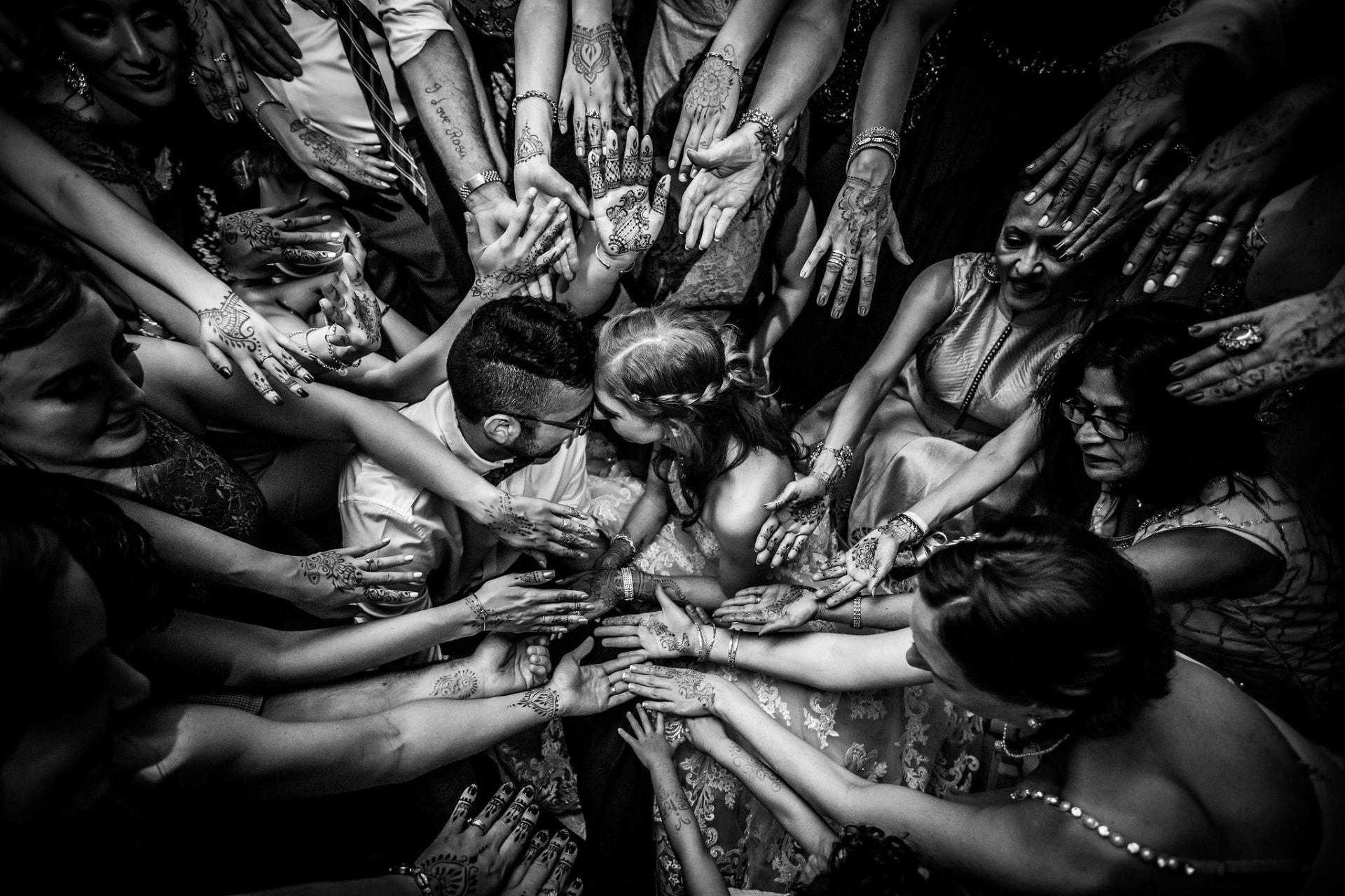 Black and white overhead photo of wedding guests reaching hennaed hands toward the couple