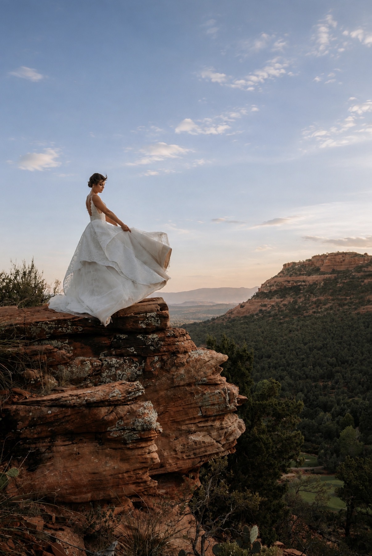 Bride holding her gown on a red rock ledge overlooking Sedona at sunset